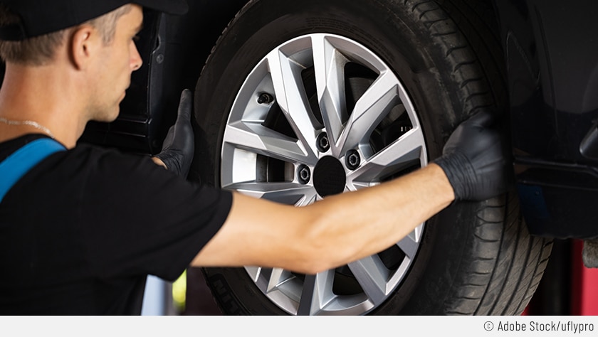 Auf dem Bild ist zu sehen, wie ein Automechaniker an einem Reifen eines auf einer Hebebühne stehenden Autos leicht rüttelt. So kann er das Radlager prüfen und einen Defekt ausschließen.