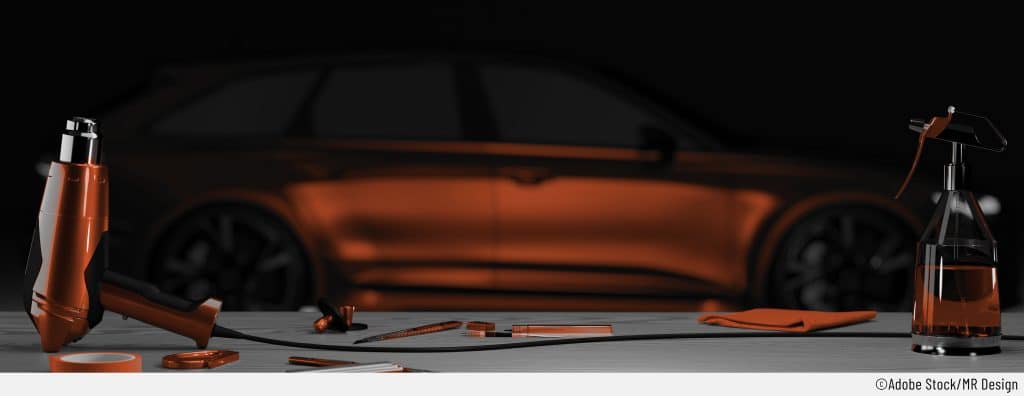 Auf dem Bild ist im Hintergrund verschwommen ein frisch in orange foliertes Auto zu sehen. Im Vordergrund sieht man auf einem Tisch klar die Hilfsmittel, die zum Auto-Folieren benötigt werden: Ein Föhn, Klebeband, Cutter-Messer, Sprühflasche mit passendem Reinigungsmittel. Das ganze Bild hat orangfarbene Akzente.