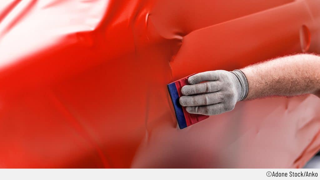 Beim Auto-Folieren wird mit einem Rakel gearbeitet, damit keine Bläschen unter der Autofolie entstehen. Auf dem Bild bekommt das Auto eine rote Folie und der Fokus liegt auf der Hand, welche Handschuhe trägt und mit einem Rakel arbeitet, um die Folie anzubringen.