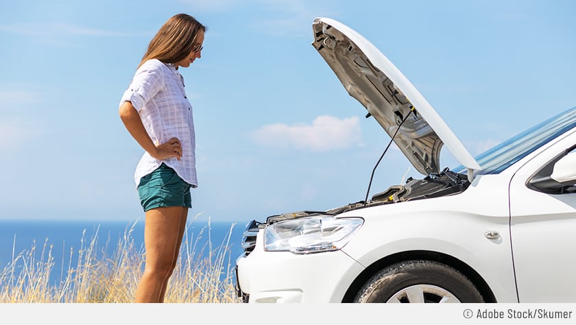 Eine Frau steht neben ihrem weißen Auto. Die Motorhaube ist geöffnet und die Frau schaut verzweifelt, ob alle Flüssigkeiten aufgefüllt sind.