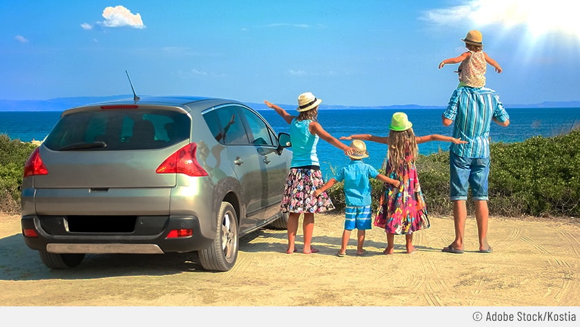 Eine fünfköpfige Familie mach Urlaub mit Auto und Kindern. Die Leute stehen neben dem grauen Pkw am Strand. Im Hintergrund sieht man blauen Himmel mit wenigen Wolken und das Meer.