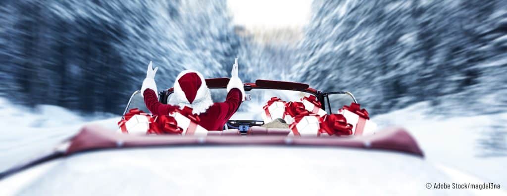 Der Weihnachtsmann sitzt mit gehobenen Händen in einem mit Geschenken voll gepackten roten Cabrio und fährt auf einer von Bäumen umgebenden verschneiten Straße.