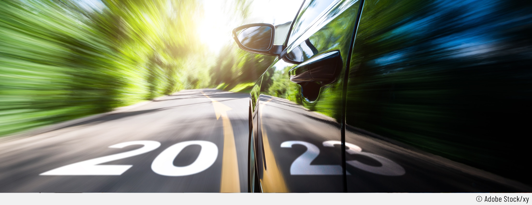 Auf dem Bild ist ein schnell fahrendes Auto aus einer tiefen Perspektive zu sehen. Der Hintergrund ist zum Darstellen der Geschwindigkeit verzerrt. Auf der Straße ist eine Markierung mit "20" und die Spiegelung im Auto ergänzt das neue Jahr mit "23".