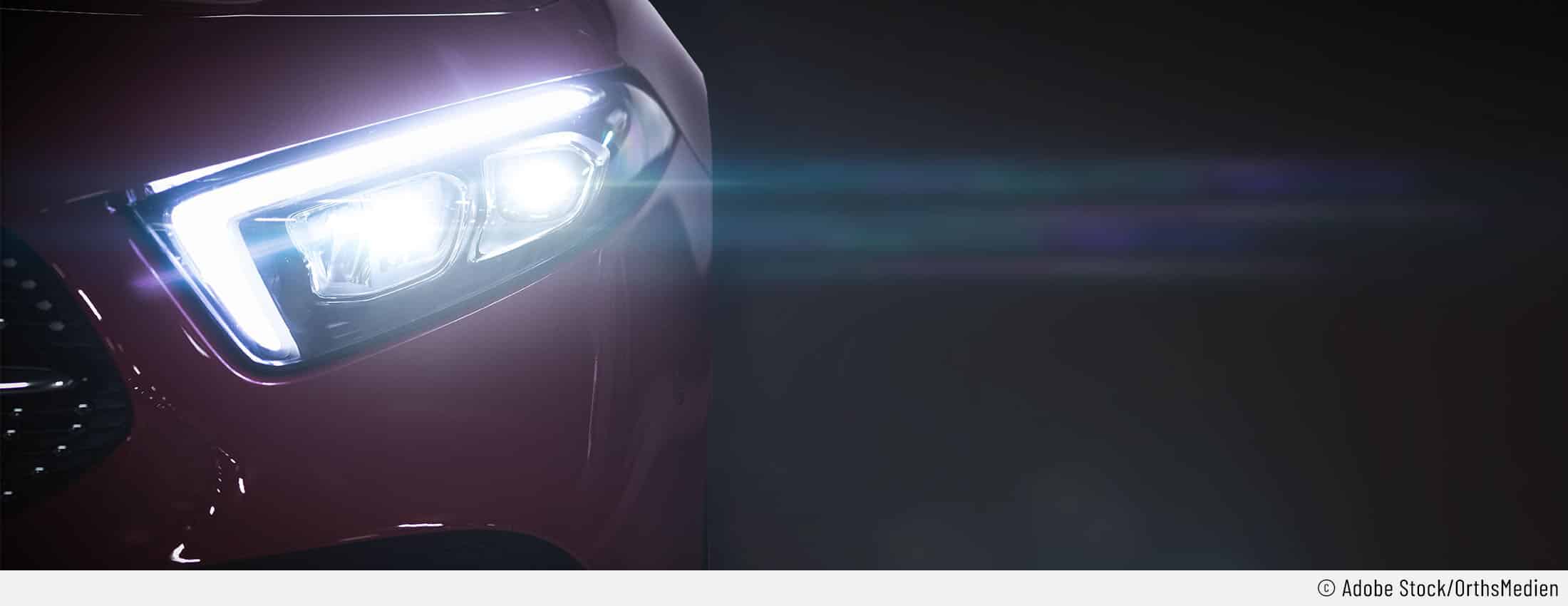 Auf diesem Bild ist ein rotes Auto in der Dunkelheit zu sehen. Der Fokus liegt auf dem linken Fernlicht, das eingeschaltet ist.