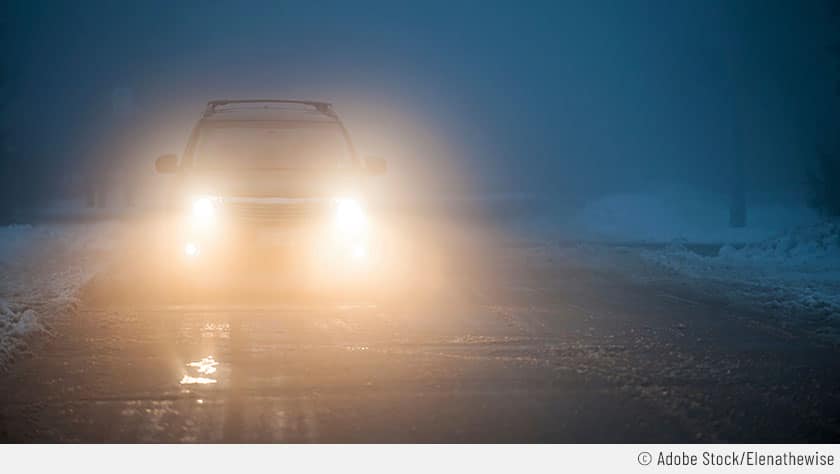 Hier sieht man ein herannahendes Auto mit eingeschaltetem Fernlicht. Links und rechts liegt Schnee auf der Straße. Es ist nebelig und deswegen blendet das Fernlicht.