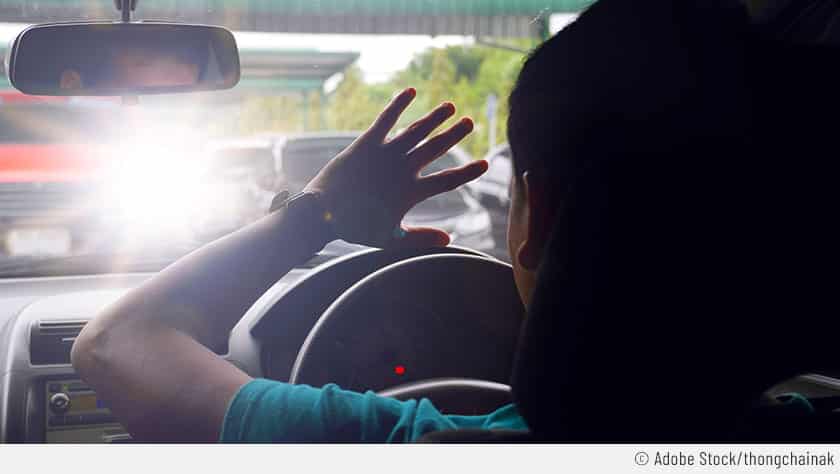 Eine Frau sitzt im Auto und streckt ihre Hand aus, um sich vor dem blendenden Fernlicht eines Fahrzeugs aus dem Gegenverkehr zu schützen.