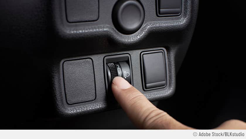 Zu sehen ist eine Nahaufnahme eines Ausschnitts des Armaturenbretts eines Autos, bei dem eine Person mit ihrem Finger an dem Rädchen zur Leuchtweitenregulierung dreht