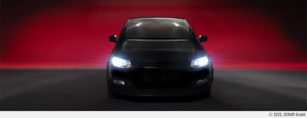 Auf dem Headerbild ist ein Auto mit eingeschalteten Frontscheinwerfer von vorne zu sehen. Es ist eindeutig mit Retrofit-LEDs ausgerüstet. Der Untergrund ist dunkelgrau. Der Hintergrund ist rötlich. Somit kommt das Scheinwerferlicht besonders zur Geltung.