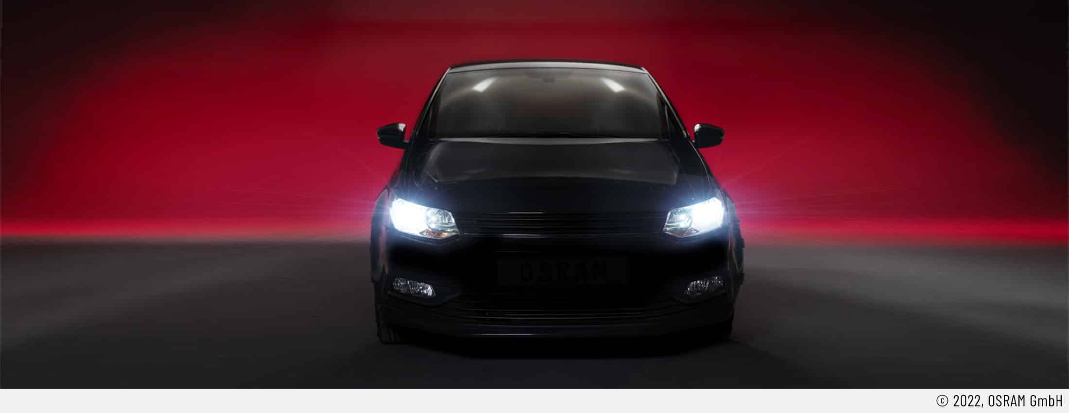 Auf dem Headerbild ist ein Auto mit eingeschalteten Frontscheinwerfer von vorne zu sehen. Es ist eindeutig mit Retrofit-LEDs ausgerüstet. Der Untergrund ist dunkelgrau. Der Hintergrund ist rötlich. Somit kommt das Scheinwerferlicht besonders zur Geltung.