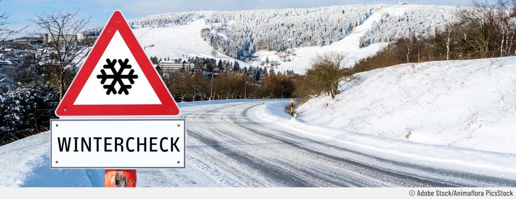 Auf dem Bild sieht man eine schneebedeckte Straße und Landschaft mit einem Verkehrszeichen im Vordergrund, das auf den Wintercheck hinweist, um das Auto winterfit zu machen.