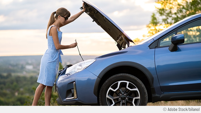 Eine Frau in einem hellblauen Sommerkleid steht vor der geöffneten Motorhaube eines silberblauen Fahrzeugs. Mit der linken Hand hält sie die Klappe oben, mit der rechten Hand hält sie den Stab, um die geöffnete Motorhaube zu fixieren.