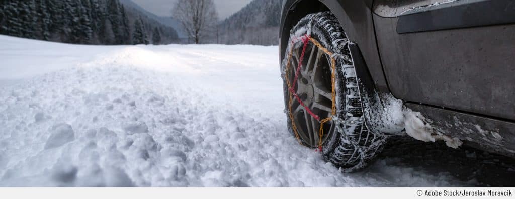 Ein Auto steht auf auf einer verschneiten Landstraße. Der Fokus liegt auf dem linken Vorderrad, das mit montierten Schneeketten daherkommt.