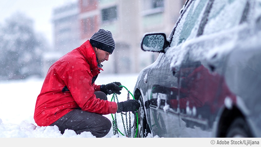 Ein Mann mit roter Winterjacke und gestreifter Mütze kniet vor dem linken Vorderrad eines silberfarbenen Autos, mit einer Reifen-Kette in der Hand. Es liegt sehr viel Schnee auf der Straße und es schneit weiterhin stark. Er möchte nun die Schneekette anbringen.