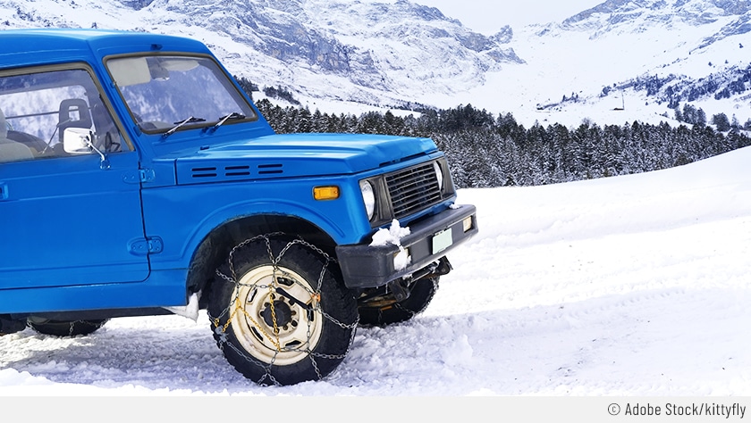 Vor einer üppig verschneiten Gebirgslandschaft steht ein blauer Geländewagen mit aufgezogenen Ketten auf allen vier Rädern.