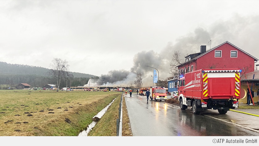 Auf einer nassen Fahrbahn stehen mehrere Feuerwehrwagen im Einsatz. Im Hintergrund steigen starke Rauchwolken aus dem brennenden Lager.