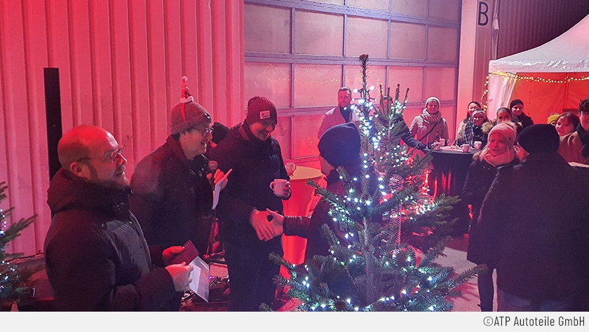 Vor der Logistik-Halle in Pressath wurden Tische und ein geschmückter Weihnachtsbaum aufgestellt. Im Hintergrund sind ATP-Mitarbeiterinnen und -Mitarbeiter zu sehen. Im Vordergrund sind die Geschäftsführer Frank Landgraf, Marc Leimann und Alexander Bugge neben dem Tannenbaum zu sehen. Sie überreichen gerade einem Jubilar ein kleines Geschenk und wirken dabei sehr fröhlich.