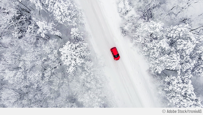 Das zu sehende Bild ist aus der Vogelperspektive aufgenommen. Darauf sieht man ein verschneites Waldstück, das von einer Straße durchzogen wird. Darauf fährt ein rotes Auto.