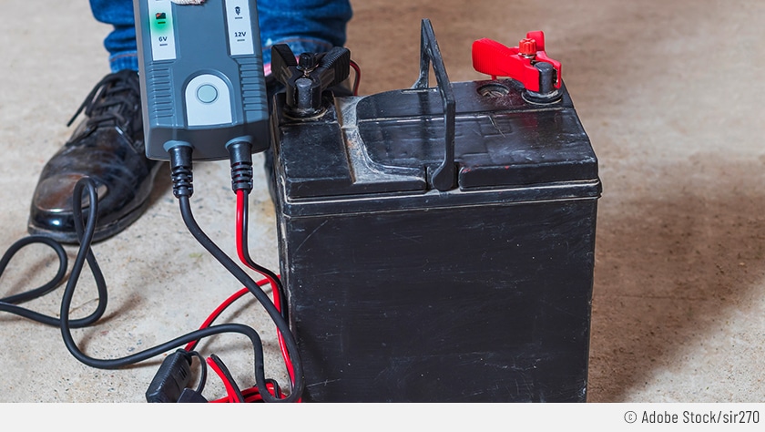 Die ausgebaute Batterie steht auf dem Boden und ist bereits an das Ladegerät angeschlossen. Der Mechaniker hält das Ladegerät in der Hand und der Fokus liegt auf der rot leuchtenden Anzeige: Er wird diese EFB-Batterie laden müssen.