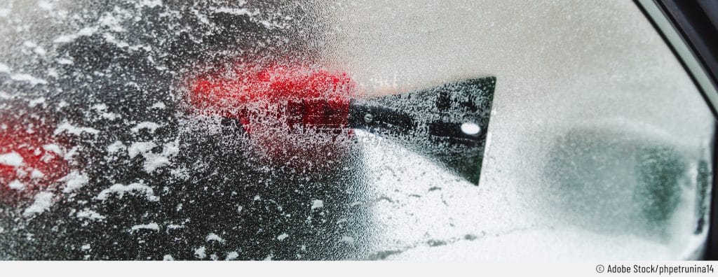 Auf dem Bild sieht man jemanden beim Eiskratzen am Auto. Die Perspektive ist innerhalb des Autos und zeigt die Person beim Enteisen der Autoscheiben von außen.