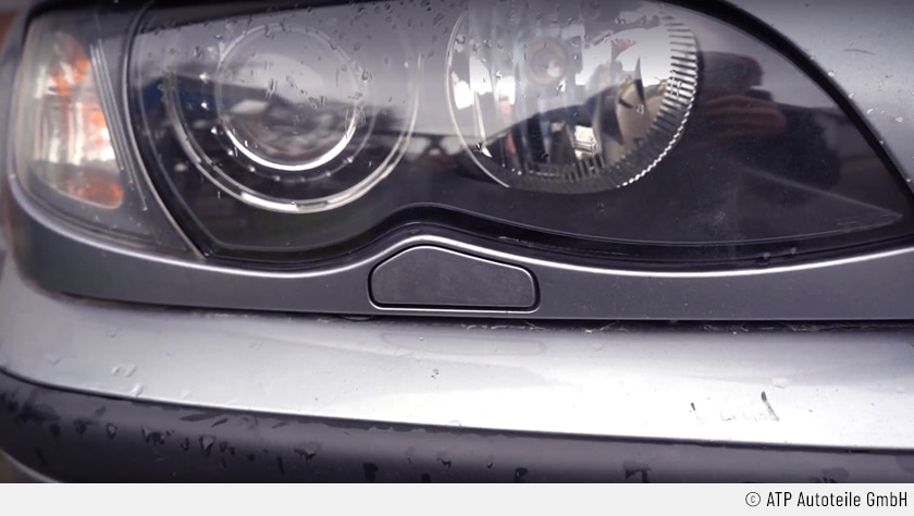 Der Fokus liegt auf dem rechten Frontscheinwerfer und der darunter zu sehenden Blende der Scheibenwischerreinigungsanlage des silberfarbenen 3er BMW E46.