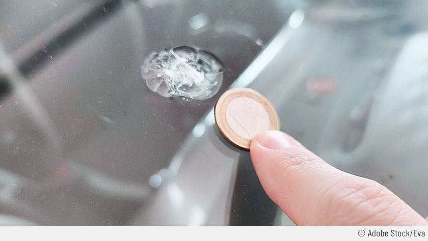 Auf dem Bild ist zu sehen, wie jemand mit einem Finger eine Münze neben den Steinschlag hält, um die Größe zu vergleichen. Dadurch kann man erkennen, ob eine Steinschlag-Reparatur zulässig ist oder nicht.