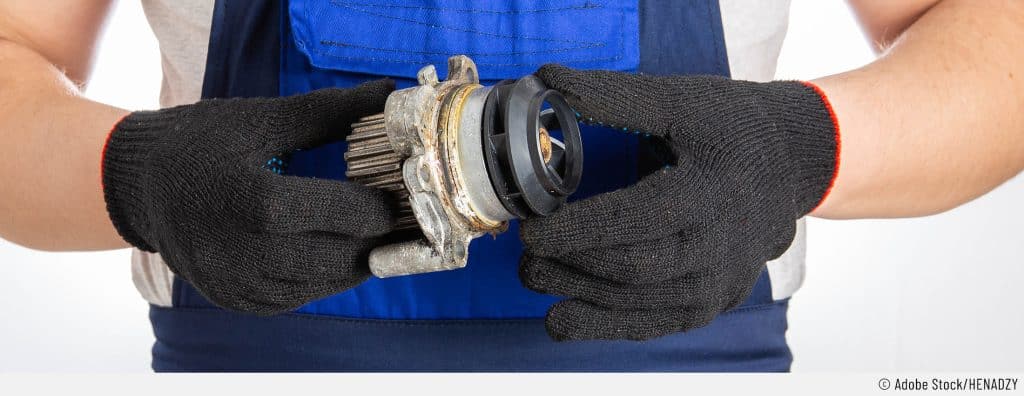 Ein Automechaniker hält eine defekte Wasserpumpe mit Handschuhe fest auf Bauchhöhe.