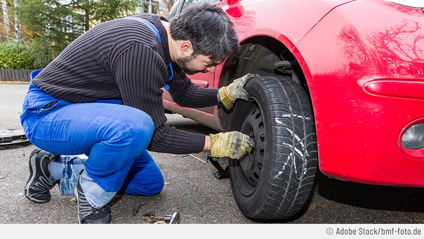 Ein Kfz-Mechaniker ist dabei, die Radschrauben bei einem roten Auto mit der Hand festzuziehen. Der Reifen ist mit "VR" beschriftet, um beim nächsten Reifenwechsel zu wissen, wo der Reifen montiert werden muss. Neben dem Mann liegt ein Drehmomentschlüssel, um die Schrauben anschließend festzuziehen.