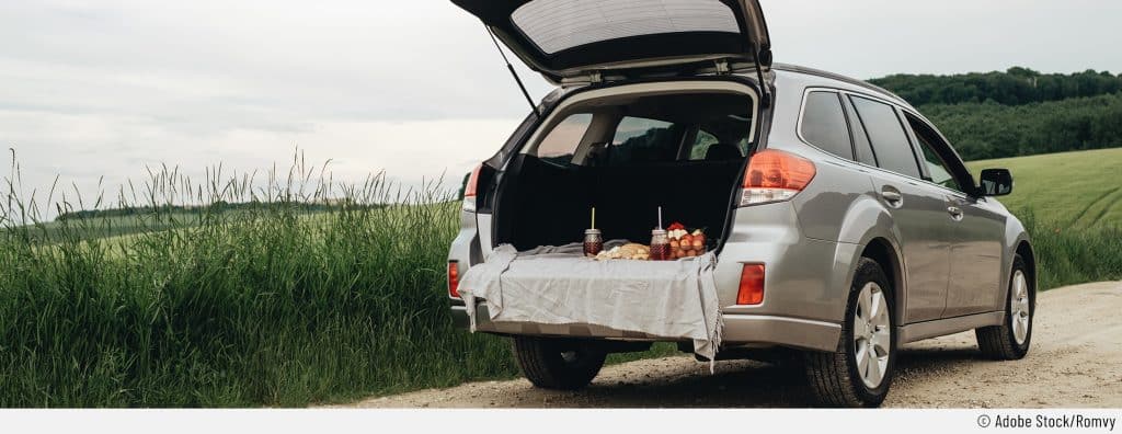 Auf dem Bild ist ein graues Auto von hinten zu sehen. Der Kofferraum des Kombis ist geöffnet. Darin ist eine Decke mit Getränken und Snack zu sehen – perfekt für ein Picknick im Auto.