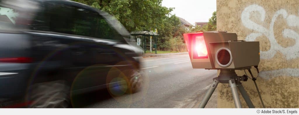 Auf dem Bild sieht man, wie ein Auto gerade geblitzt wird, weil die Person zu schnell gefahren ist. Rechts ist der stationäre Blitzer mit rotem Licht zu sehen. Links verschwommen ein schnell vorbeifahrendes Auto.