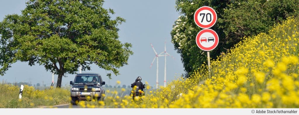 Auf einer Landstraße fahren ein SUV und ein Motorrad durch eine Zone mit Überholverbot. Durch Verkehrsschilder wird die Geschwindigkeit auf 70 km/h beschränkt und das Überholen für Fahrzeuge aller Art verboten. Sowohl links als auch rechts im Hintergrund sind Bäume zu sehen. Im Vordergrund sind gelbe Blumen, jedoch liegt der Fokus auf den Verkehrszeichen.