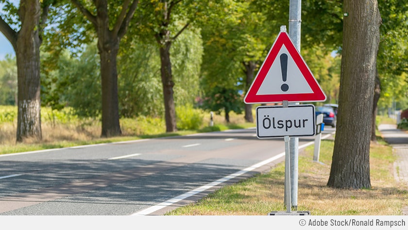 An einer Landstraße ist ein Ölspur-Schild zu sehen. In dem Fall wird die Ölspur mit der Kombination Gefahrenzeichen plus Schriftzug angezeigt.