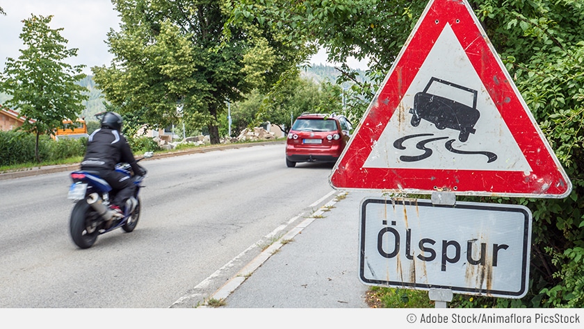 An einer befahrenen Landstraße ist ein Ölspurschild zu sehen. Dieses besteht aus einem Sinnbild (mit einem ins Schleudern geratenen Auto) und dem eindeutigen Schriftzug.