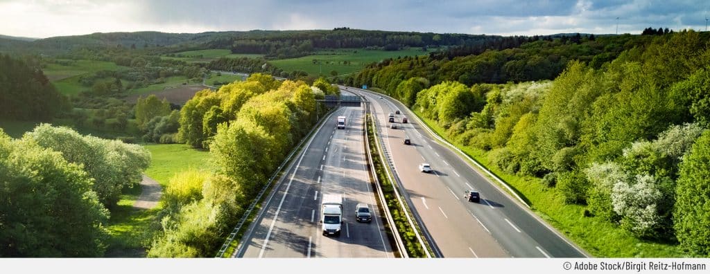 Zu sehen ist ein Teilabschnitt einer Autobahn. Rund herum ist viel Grün zusehen: Büsche, Bäume und Wiese umranden die Straße. Dies dient als Symbolbild für lange Autofahrten.