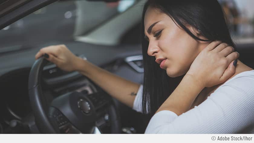 Zu sehen ist eine Frau im Auto mit schmezerfülltem Blick nach einer langen Autofahrt. Eine Hand liegt auf dem Lenkrad, während die andere den Nacken hält.