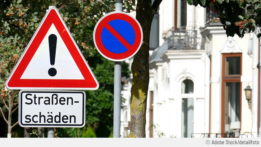In der Mitte des Bildes ist das Verkehrsschild  für eingeschränktes Halteverbot zu sehen. Im Hintergrund ist ein Haus zu erkennen und links im Bild ein Schild, das auf Straßenschäden hinweist.