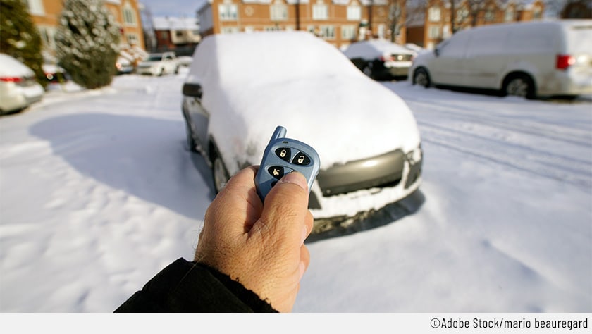 In einer schneebedeckten Landschaft hält ein Mann einen elektronischen Schlüssel in der Hand, anhand dessen er die Standheizung aus der Ferne aktivieren kann.  Das ebenfalls schneebedeckte Auto dürfte damit den bevorstehenden Kaltstart besser überwinden.