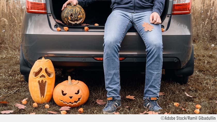 Halloween-Auto mit offenem Kofferraum. Neben der darauf sitzenden Person sind drei geschnitzte Kürbisse zu sehen.